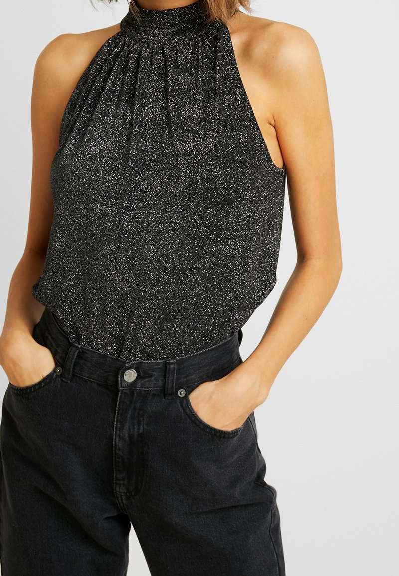 Musta halterneck-toppi, jossa on rypytetty kaulussuunnittelu ja kimaltava pinta. Yhdistetty korkeavyötäröisiin tummiin farkkuihin, kädet taskuissa.