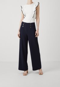 White top with ruffled sleeves and black trim, paired with navy wide-leg trousers featuring gold buttons. Smooth texture throughout.