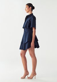 Tamsiai mėlyna suknelė su apykakle, trumpomis rankovėmis ir sluoksniuotais volanais. Pora su kreminės spalvos aukštakulniais sandalais ant juostelių. Lygus paviršius.