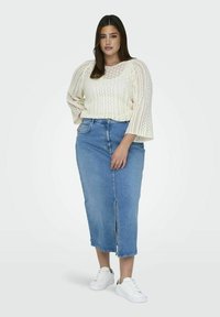 Cremfarbener gestrickter Pullover mit weiten Ärmeln, kombiniert mit einem hellblauen Denimrock mit einem Vorder-Schlitz, ergänzt durch weiße Sneakers.