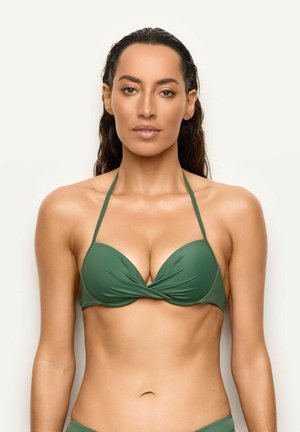 Donna con lunghi capelli scuri che indossa un top bikini verde halter, rivolta in avanti su uno sfondo chiaro e uniforme.