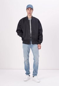 Black bomber jacket with a zipper, worn over a gray shirt. Light blue jeans and white sneakers complete the outfit. Light blue cap on the head.