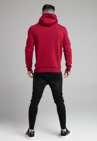 Rode katoenen hoodie met een grote capuchon, strakke mouwen en een geribbelde zoom, gecombineerd met zwarte skinny jeans en zwarte schoenen met gouden accenten.