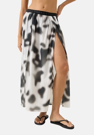 Kobieta ubrana w długą spódnicę w czarno-biały wzór z rozcięciem do uda, czarnym paskiem w talii i czarnymi klapkami, stojąca z jedną ręką na biodrze.