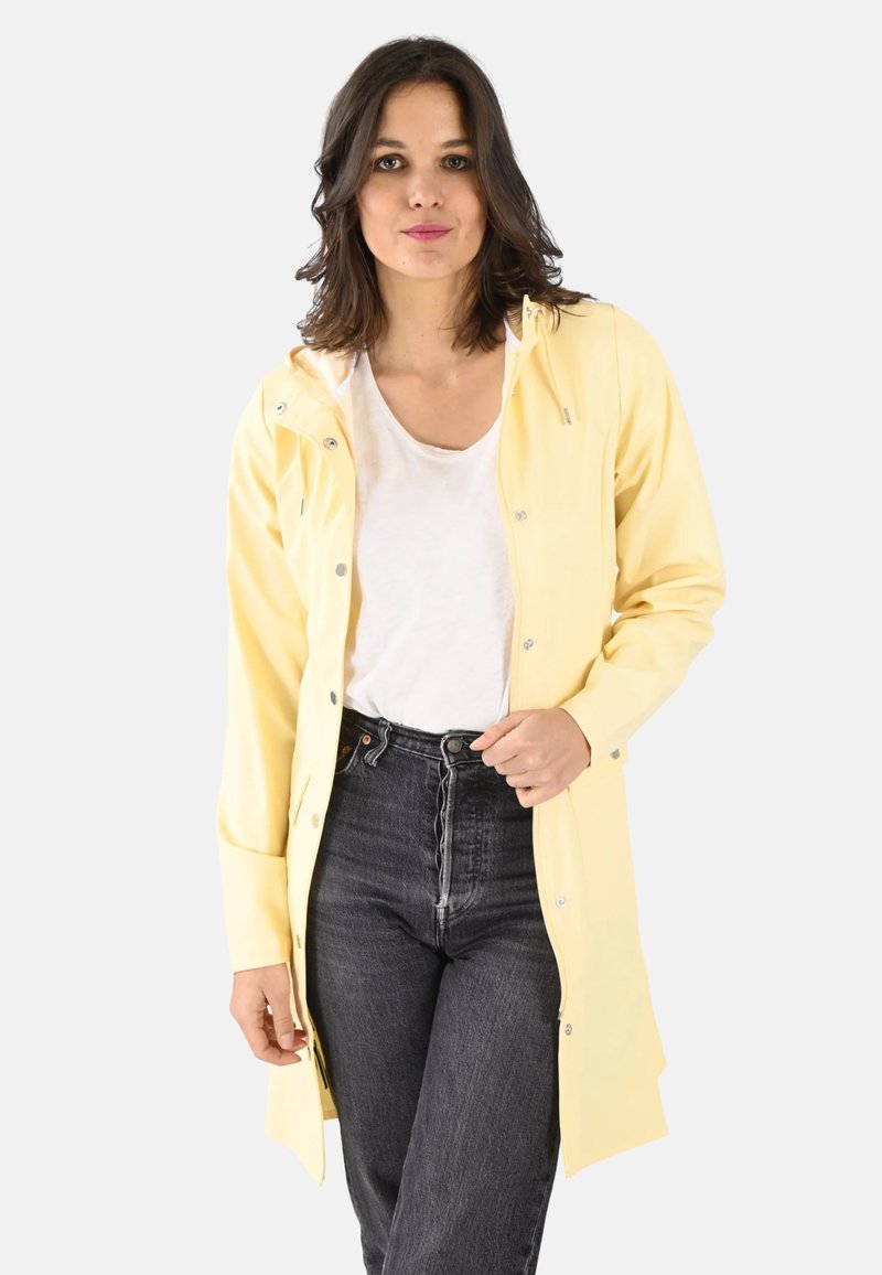 Veste imperméable jaune avec capuche, dotée de boutons-pression, de manches longues et d'une coupe droite, associée à un jean noir taille haute.