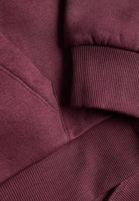 Gros plan d’un sweat-shirt bordeaux montrant le poignet côtelé, l’ourlet et une partie d’une poche kangourou avec des coutures visibles.