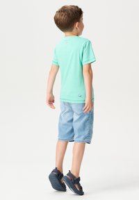Minzgrünes T-Shirt mit kurzen Ärmeln, blaue Jeansshorts und dunkle Sandalen. Das T-Shirt hat ein Etikett auf der Rückseite und dezente Nähte.