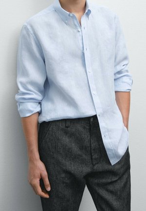 Homme portant une chemise bleue claire à manches longues retroussées et un pantalon gris foncé, une main dans la poche, sur fond uni.