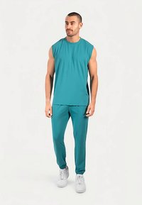 Turquoise mouwloze top en bijpassende broek van gladde stof. De outfit heeft een ontspannen pasvorm en is gecombineerd met witte sneakers.