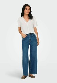 Vit blus med vita prickar och V-ringning, kombinerad med högmidjade vida blå jeans. Modellen bär bruna ballerinaskor. Enkel, modern stil.