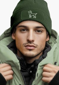 Green ribbed beanie with a dog design on a rectangular patch, worn with a light green puffer jacket layered over a black inner jacket.