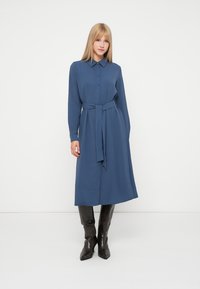 Kék, hosszú ujjú ruha inggallérral, elülső gombokkal és derékon megkötött övvel. Magas fekete csizmával viselve. Sima szövet textúra.