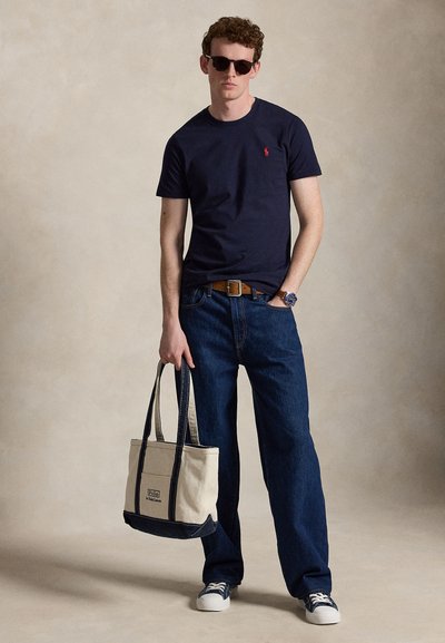 Ung mand iført solbriller, marineblå t-shirt, jeans og sneakers, der holder en beige og marineblå tote taske med den ene hånd.