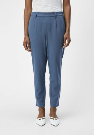 Pantaloni su misura blu con pieghe frontali, tasche laterali e gambe affusolate. Finissimo tessuto morbido, abbinati a scarpe bianche con punta a punta.
