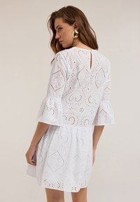Rochie albă cu broderie în puncte, mâneci evazate și detaliu cu deschidere la spate. Materialul este ușor, cu o textură subtilă.