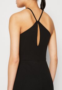 Femme en robe noire sans manches avec de fines bretelles se croisant dans le haut du dos et une petite découpe en forme de trou de serrure juste en dessous du fermoir.