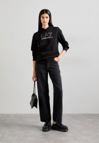 Fekete pamut kapucnis pulóver "EA7 Emporio Armani" logóval, magas derékú fekete farmernadrággal és vastag fekete cipővel, egy kis kézitáskát tartva.