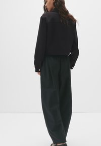Jachetă neagră scurtă cu un design structurat și mâneci lungi, asortată cu pantaloni largi de culoare verde închis, dintr-un material texturat, cu hardware minim.