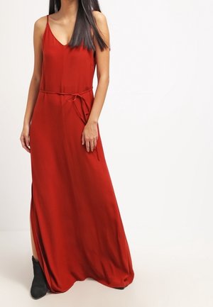 Robe maxi rouge sans manches avec fines bretelles et ceinture à la taille, portée avec des bottines noires.