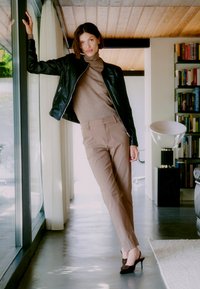 Zwarte leren jasje, bruin aansluitende coltrui, beige op maat gemaakte broek, en donkerbruine hakken; gladde texturen, eenvoudige silhouet, binnenomgeving.