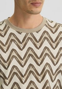 Camiseta de algodón de manga corta con una base crema, que presenta un patrón en zigzag en tonos marrones y acentos topo, con un escote redondo.