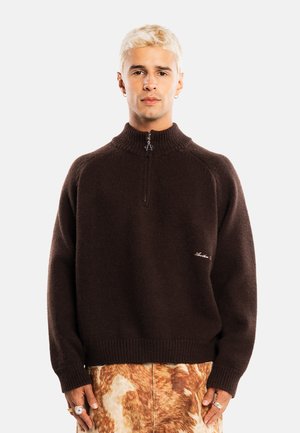 Joven con cabello rubio corto que lleva un suéter de color marrón oscuro con cremallera en la parte delantera y falda o pantalones marrones con estampado.