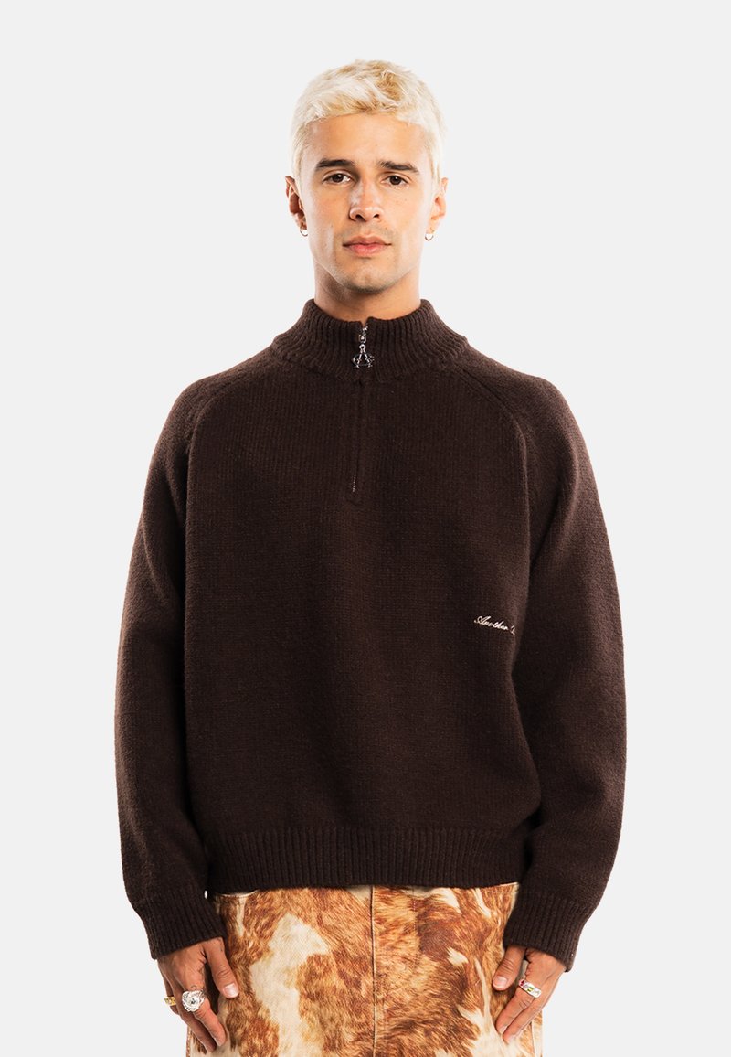 Jeune personne aux cheveux blonds courts portant un pull marron foncé avec une fermeture éclair à l'avant et une jupe ou un pantalon marron à motifs.