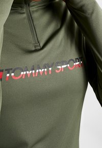 Olivegrünes sportliches Langarmshirt mit einem Viertelreißverschluss und dem "TOMMY SPORT"-Logo in Schwarz, Rot und Weiß auf der Vorderseite. Glatte Textur.