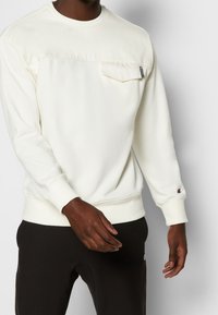 Felpa beige chiaro con scollo rotondo, tasca frontale e polsini a costine. Presenta una piccola toppa con logo sulla tasca. Indossata con pantaloni scuri.