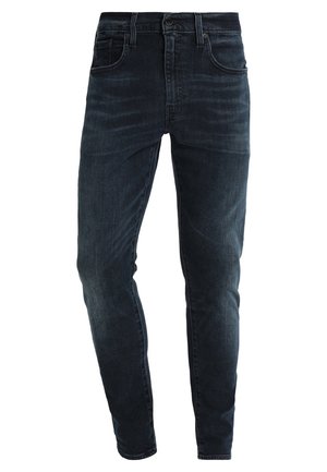 Jean slim bleu foncé avec poches avant et arrière, passants pour ceinture et fermeture par bouton.