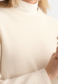 Suéter tejido de color crema con un cuello alto acanalado y superficie texturizada; mangas largas con puños acanalados.