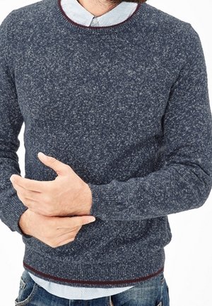 Mężczyzna ubrany w ciemnoszary melanżowy sweter na jasnobłękitnej koszuli z kołnierzykiem, zaciska lewą rękę prawą dłonią.