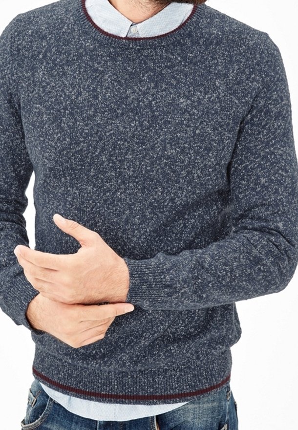 Homme portant un pull gris foncé moucheté sur une chemise bleu clair à col, serrant son poignet gauche avec sa main droite.