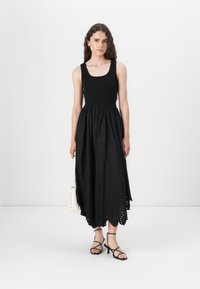 Black dress with a fitted bodice and a flared skirt featuring eyelet detailing along the hem. The model wears strappy sandals and holds a bag.