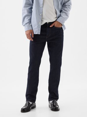 Dark blue denim jeans with a straight-leg fit, featuring five pockets, and stitched detailing, paired with a light striped button-up shirt.