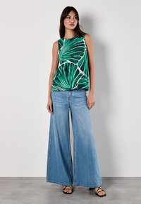 Ärmelloses grünes Oberteil mit weißem Blattmuster, kombiniert mit hellblauen weit geschnittenen Jeans und schwarzen Sandalen mit Riemchen. Glatte Stofftextur.