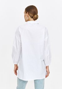 Biała, długopolska koszula z luźnym krojem i opadającymi ramionami; wykończona zaokrąglonym dołem i kołnierzykiem zapinanym na guziki. Gładka struktura materiału.