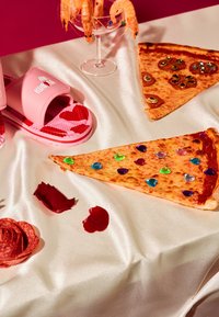 Różowe klapki z serduszkami obok dwóch kawałków pizzy ozdobionych kolorowymi kamieniami szlachetnymi, na kremowej satynowej powierzchni z płatkami róż.