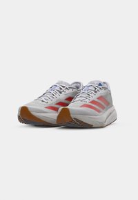 Lichtgrijze sport schoenen met rode accenten, textielen bovenkant, gevoerde kraag en rubberen loopzool met horizontale groeven voor grip.