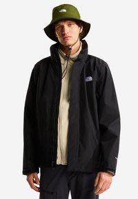 Mies, jolla vihreä kalastajanhattu leukaremmillä, beige vetoketjullinen fleece, musta vetoketjullinen takki ja mustat housut valkoista taustaa vasten.