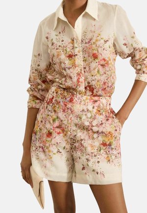 Donna che indossa una camicia crema a fiori con bottoni abbinata a pantaloncini floreali, tiene una pochette beige in una mano, l'altra mano in tasca.