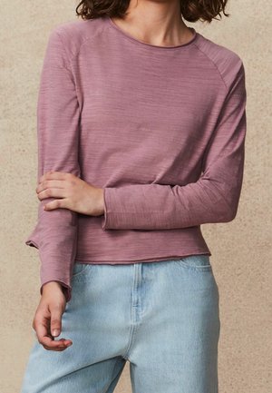 Personne portant un haut à manches longues de couleur mauve et un jean bleu clair, avec un bras croisé et l'autre reposant sur l'avant-bras.