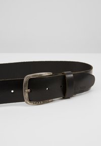 Cinturón de cuero negro que presenta una hebilla de metal en tono plateado, logo grabado y múltiples orificios para ajuste de talla. Textura suave con un acabado mate.