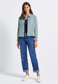 Žena stojí oblečená v světlemodré košili na knoflíky přes tmavý top, s vyhrnutými modrými džínami a bílými teniskami na jednobarevném pozadí.