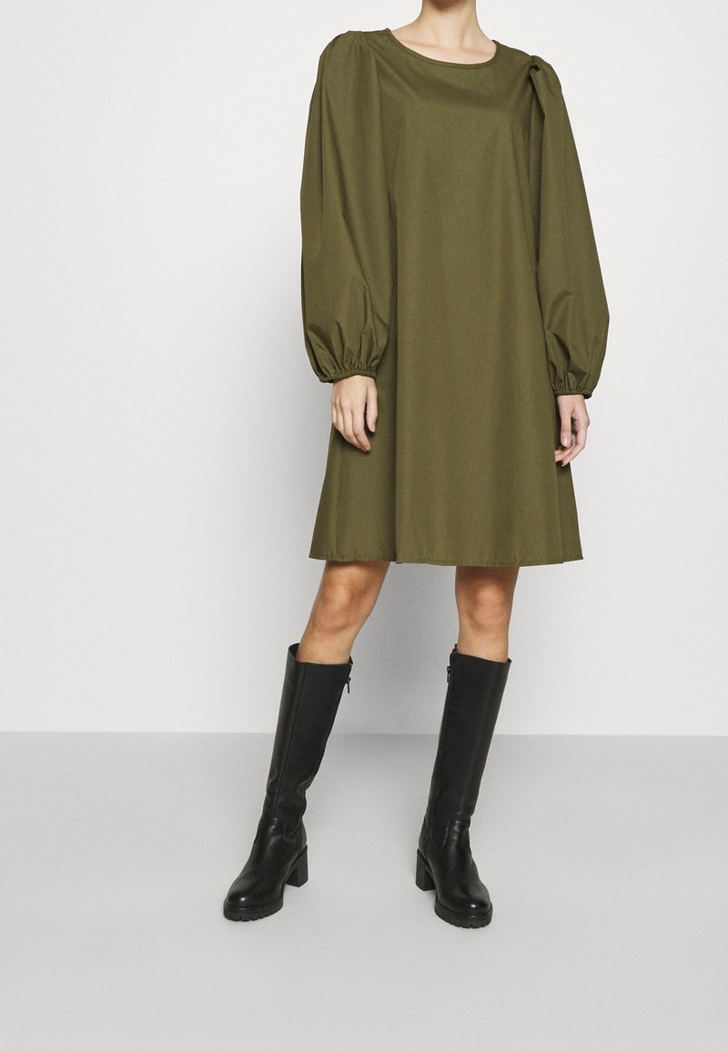 Robe vert olive aux manches longues bouffantes et col rond, confectionnée dans un tissu lisse, accompagnée de bottes noires montantes jusqu'au genou.