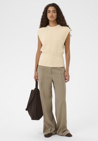 Frau mit lockigem Haar, die ein ärmelloses beiges Top und weite taupefarbene Hosen trägt, hält eine große dunkle braune Wildleder-Tasche.