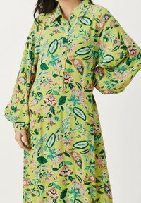 Robe florale verte avec un col, un devant à boutons et des manches amples. Le tissu présente des motifs botaniques vibrants de rose et de vert.
