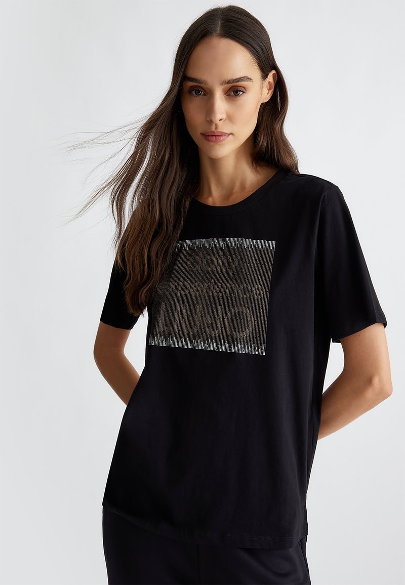 La maglietta nera presenta maniche corte e un col rotondo. La parte anteriore mostra un design grafico testurizzato in colori contrastanti.