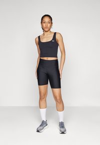 Črn športni crop top in kolesarske hlače iz raztegljivega materiala, v kombinaciji s sivimi supergami in belimi nogavicami, stoji ob preprosti podlagi.