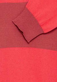Gros plan sur la manche d'un pull en maille rayé rouge et rouge foncé avec un bord-côte, reposant sur un tissu assorti en arrière-plan.